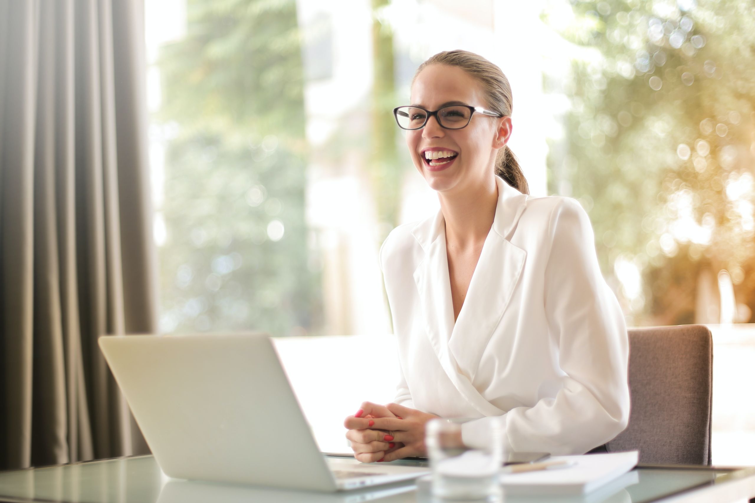 A great recruiter can significantly improve your hiring process. Blonde woman with glasses sits at a desk with a laptop and smiles.