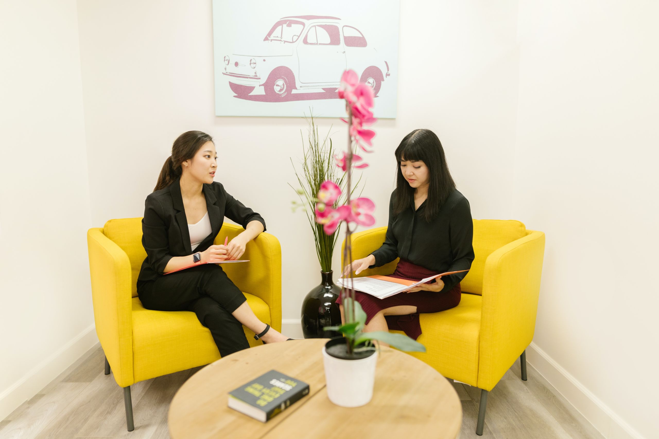 Two women sit on bright yellow chairs in an office room discussing recruiting strategies.